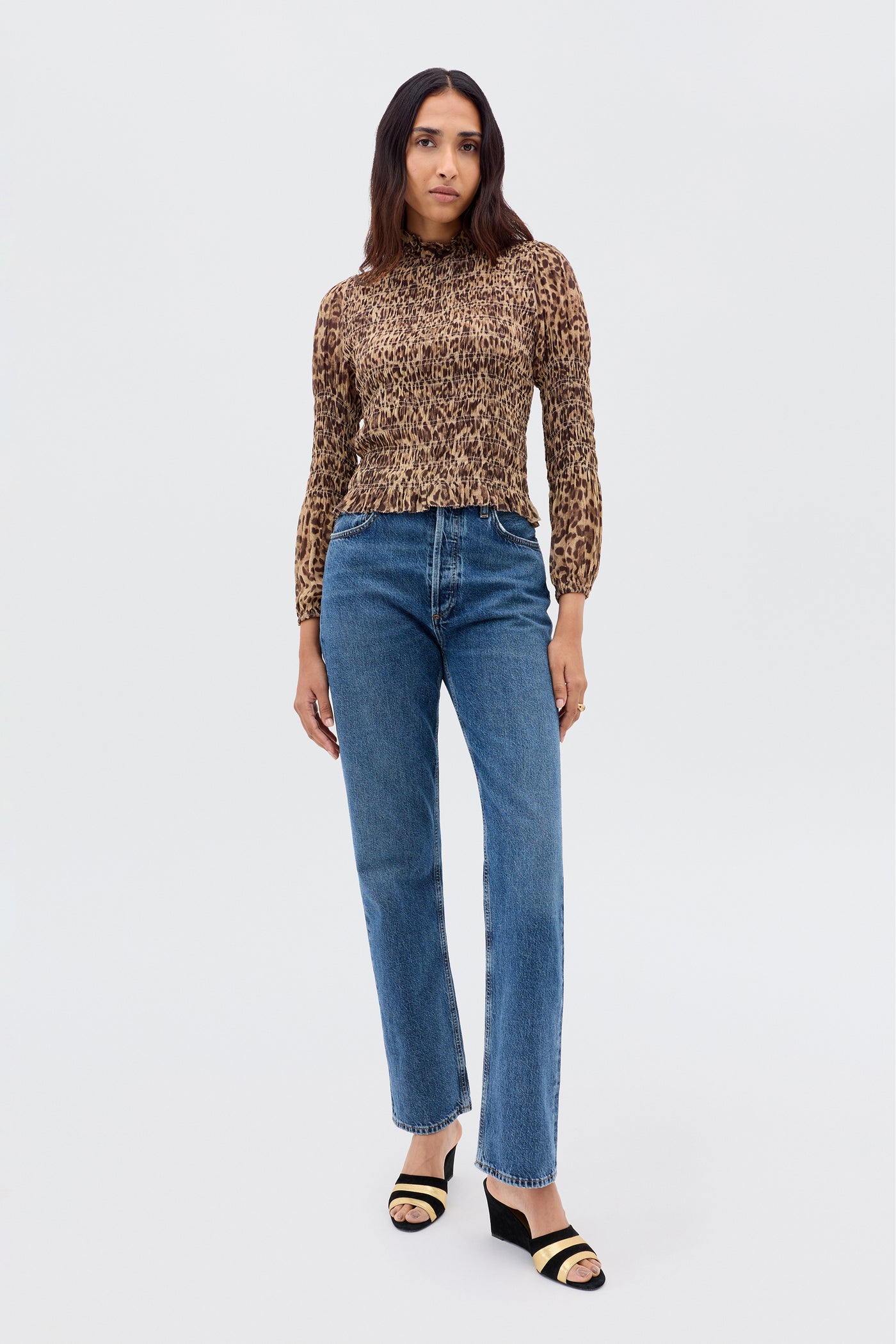 A woman with long, dark hair and a neutral expression wears the Gardenia - Mila Leopard Brown blouse with long sleeves, paired with blue high-waisted jeans and black and beige open-toe heeled sandals against a plain background.