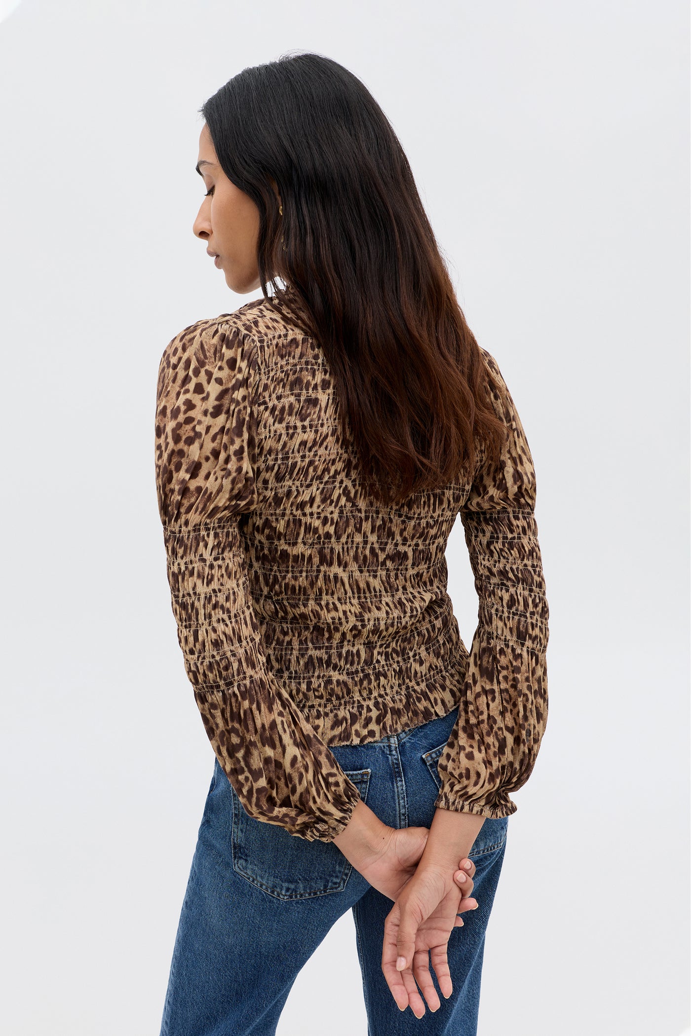 A woman with long dark hair stands with her back to the camera, wearing the Gardenia - Mila Leopard Brown blouse and blue jeans. Her hands are clasped behind her back against a plain light background.