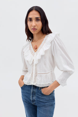 A woman with long brown hair wears the Solene - Ivory blouse—vintage-inspired with puffed sleeves, lace details, and a ruffled V-neckline—paired with blue jeans as she stands against a plain light background, hands in her pockets.