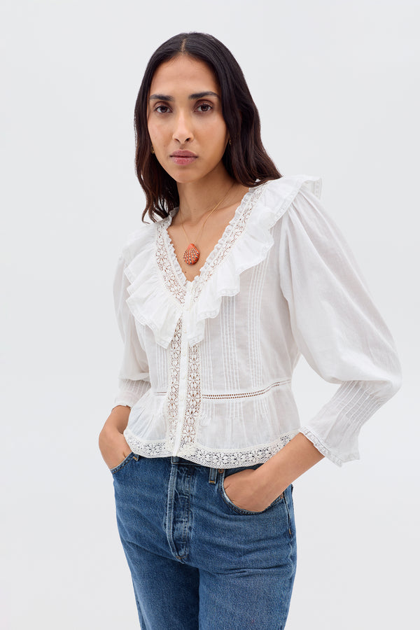 A woman with long brown hair wears the Solene - Ivory blouse—vintage-inspired with puffed sleeves, lace details, and a ruffled V-neckline—paired with blue jeans as she stands against a plain light background, hands in her pockets.