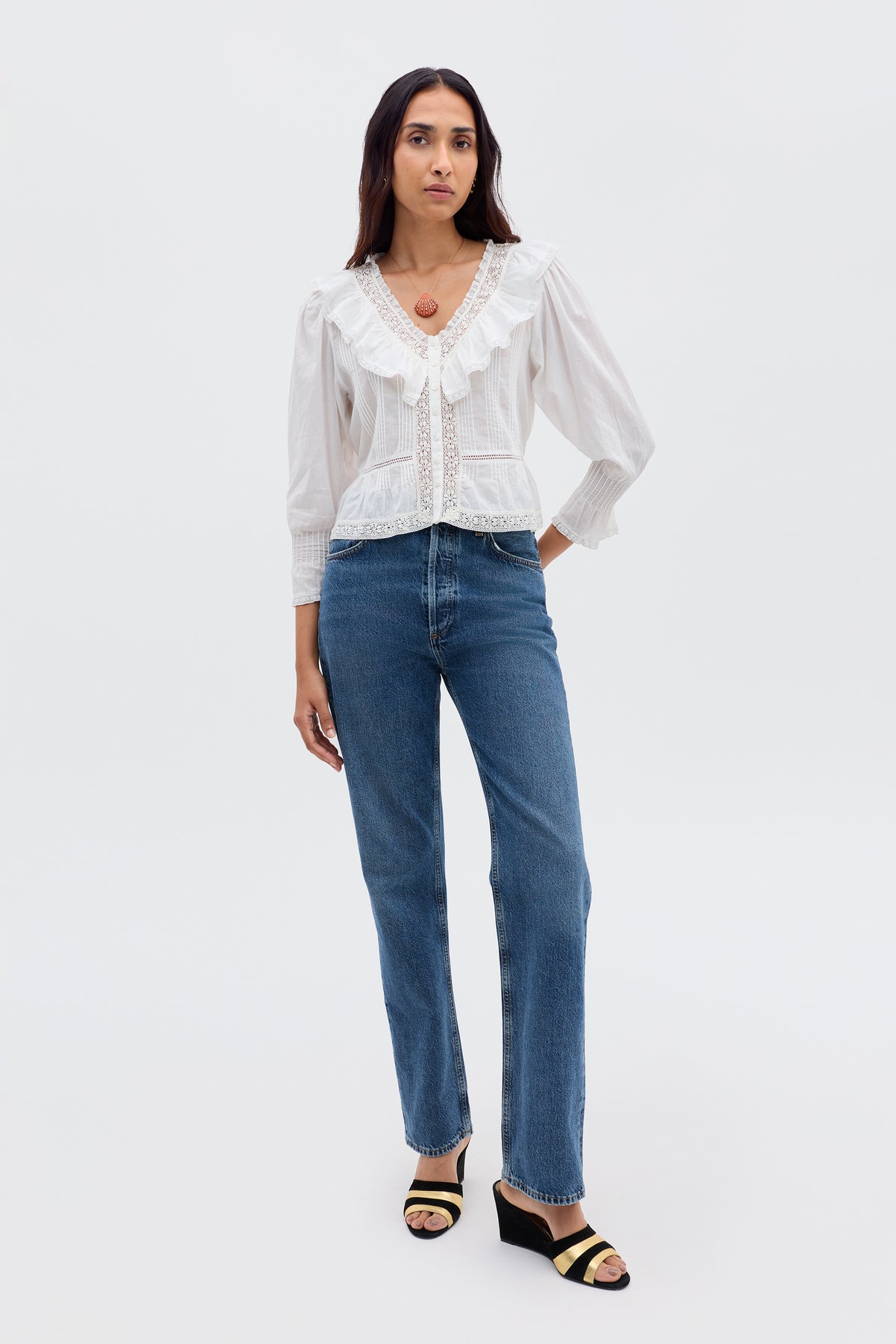A woman stands against a plain light background, wearing the Solene - Ivory vintage-inspired blouse with puffed sleeves and ruffle details, paired with blue straight-leg jeans, black heeled sandals, and a round pendant necklace.