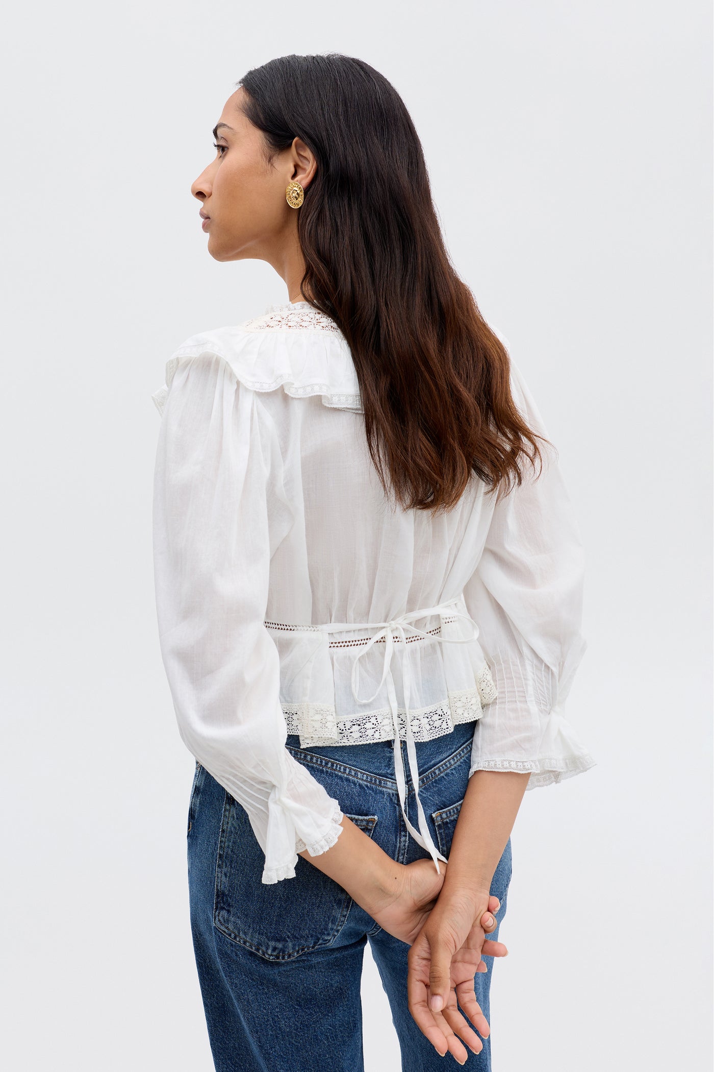 A woman with long brown hair stands facing away, wearing the Solene - Ivory blouse with puffed sleeves and a ruffled V-neckline, paired with blue jeans. Her hands are clasped behind her back.