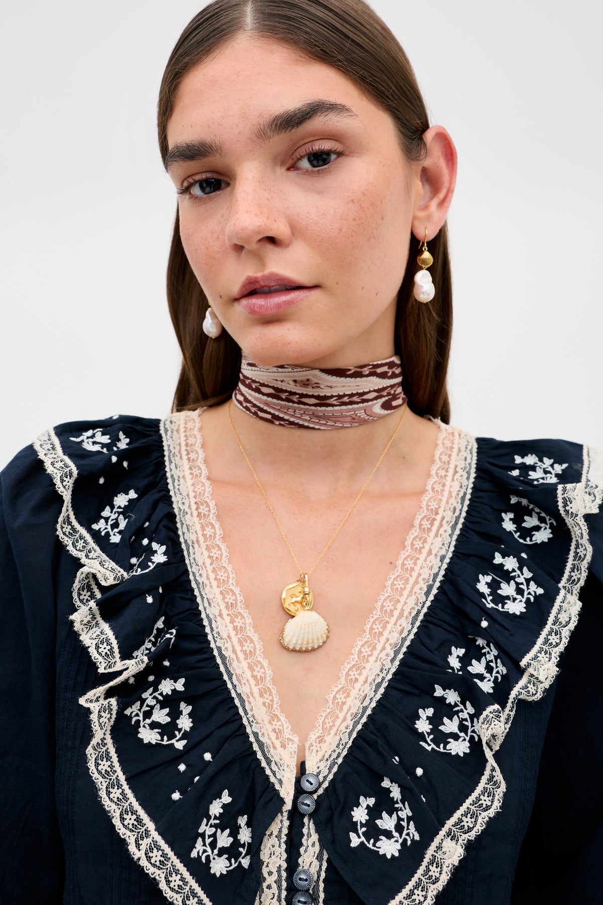 Woman with straight brown hair wearing a black blouse with lace and floral embroidery, a patterned choker, vintage jewellery including pearl earrings, and layered gold necklaces—one featuring a Devine necklace with a coin pendant and the other adorned with the Shell - White gold-plated shell charm—posing against a plain background.