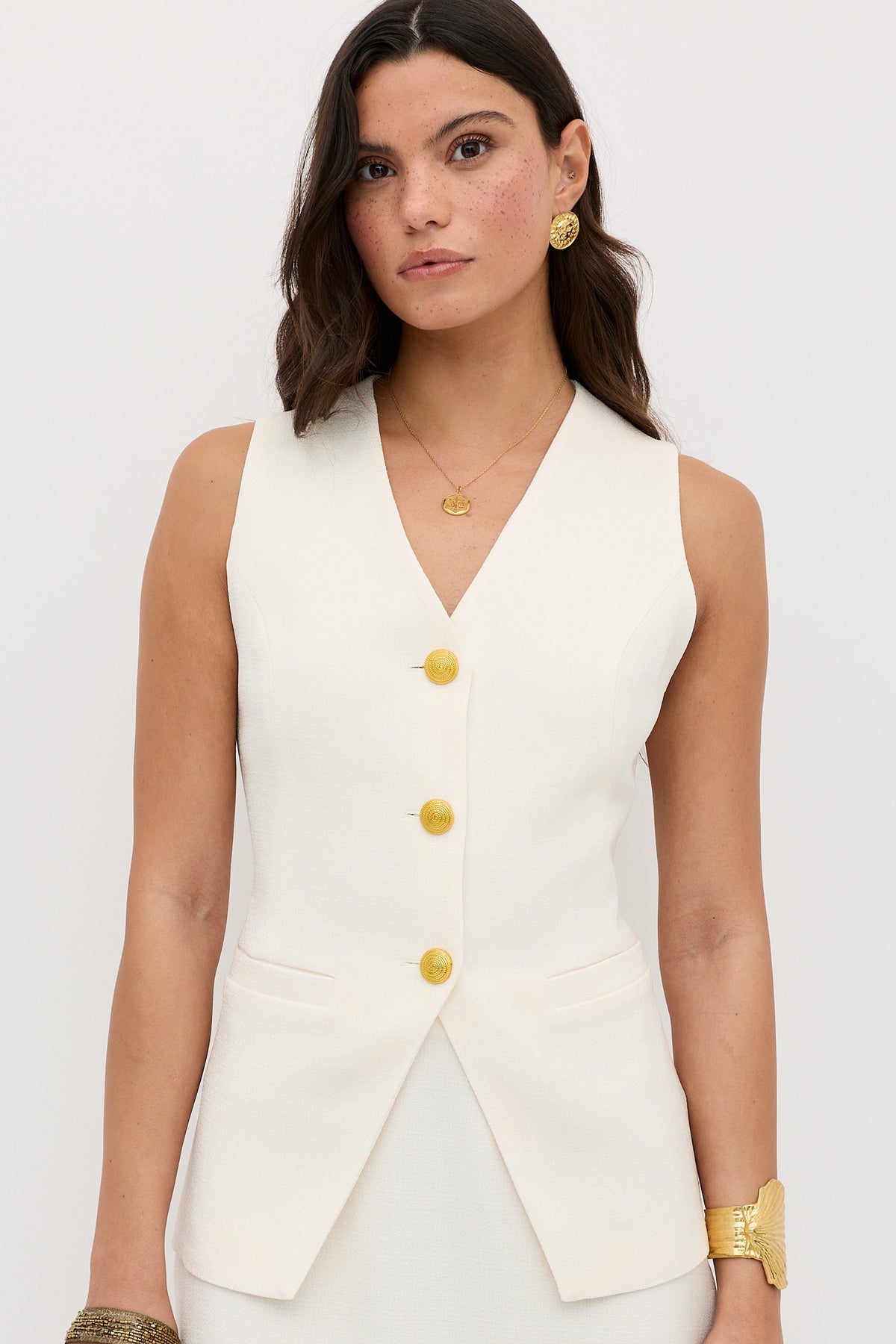 A woman with wavy brown hair wears the Ingrida - Pearl sleeveless white vest with gold buttons and gold jewelry, standing against a plain light background.