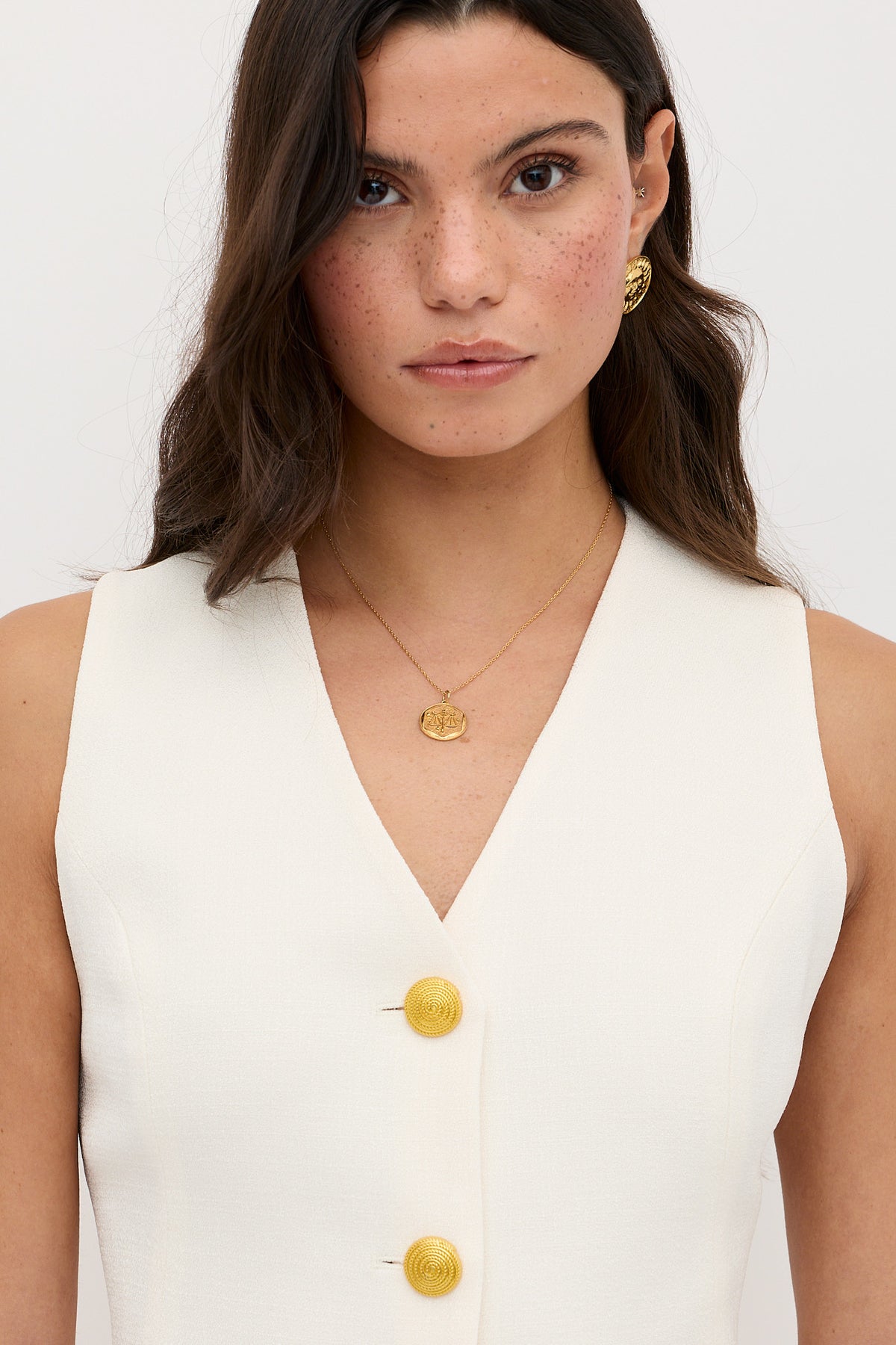 A woman with long brown hair and freckles wears the Ingrida - Pearl sleeveless white top with large yellow buttons, gold earrings, and a gold pendant necklace. She looks directly at the camera against a plain white background.
