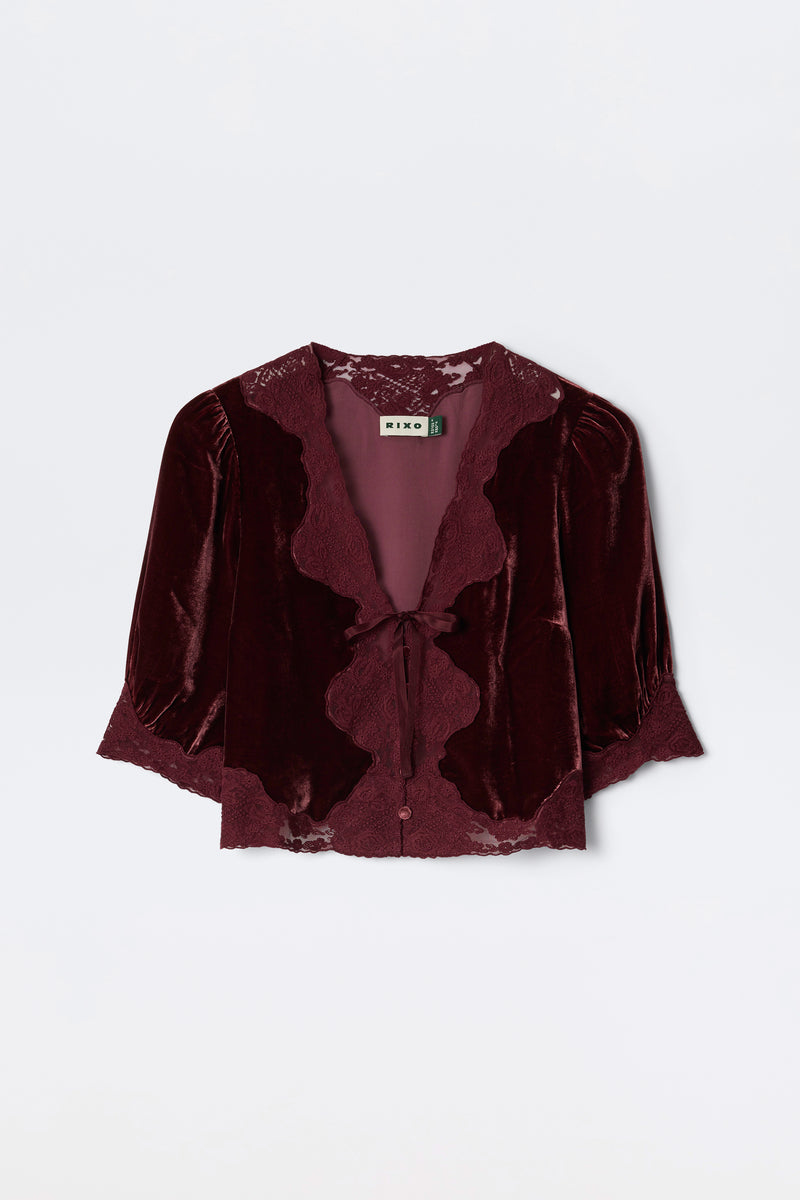 The Ursula - Bordeaux is a burgundy silk-velvet blouse featuring lace trim, puffed mid-length sleeves, and a front tie closure. Vintage-inspired, it’s finished with a “RIXO” label at the neckline.