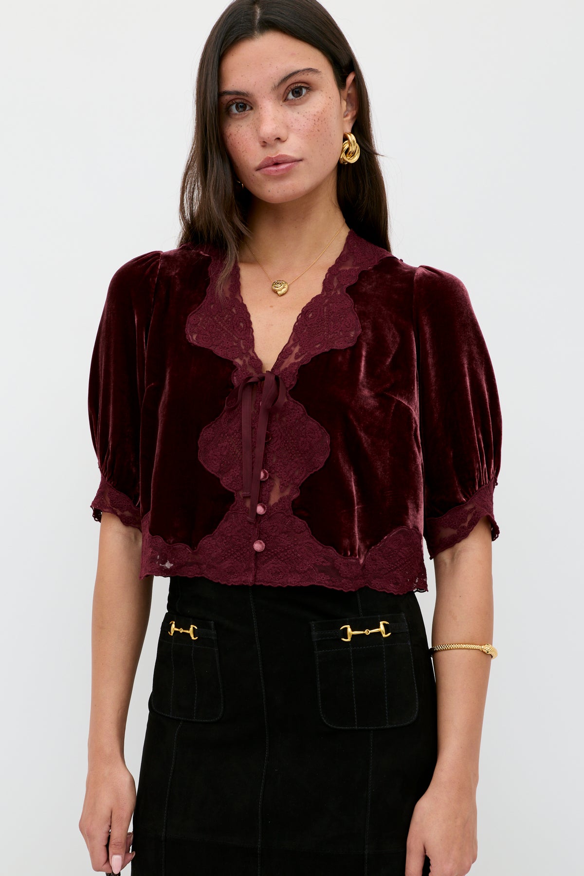 A woman with long dark hair and gold jewelry wears the Ursula - Bordeaux vintage-inspired silk-velvet blouse with lace trim and neck bow, paired with a black skirt featuring gold-accented front pockets, standing against a light background.
