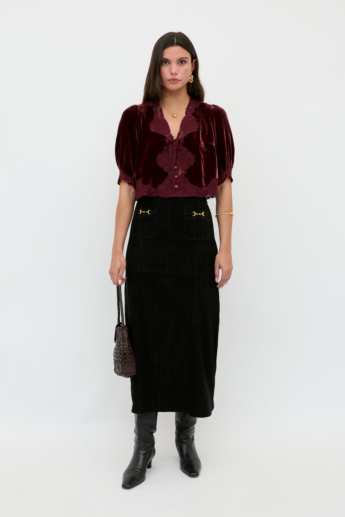 A woman stands against a white backdrop wearing the Ursula - Bordeaux, a vintage-inspired maroon silk-velvet blouse with puff sleeves, styled with a long black skirt, knee-high boots, gold jewelry, and a textured handbag.