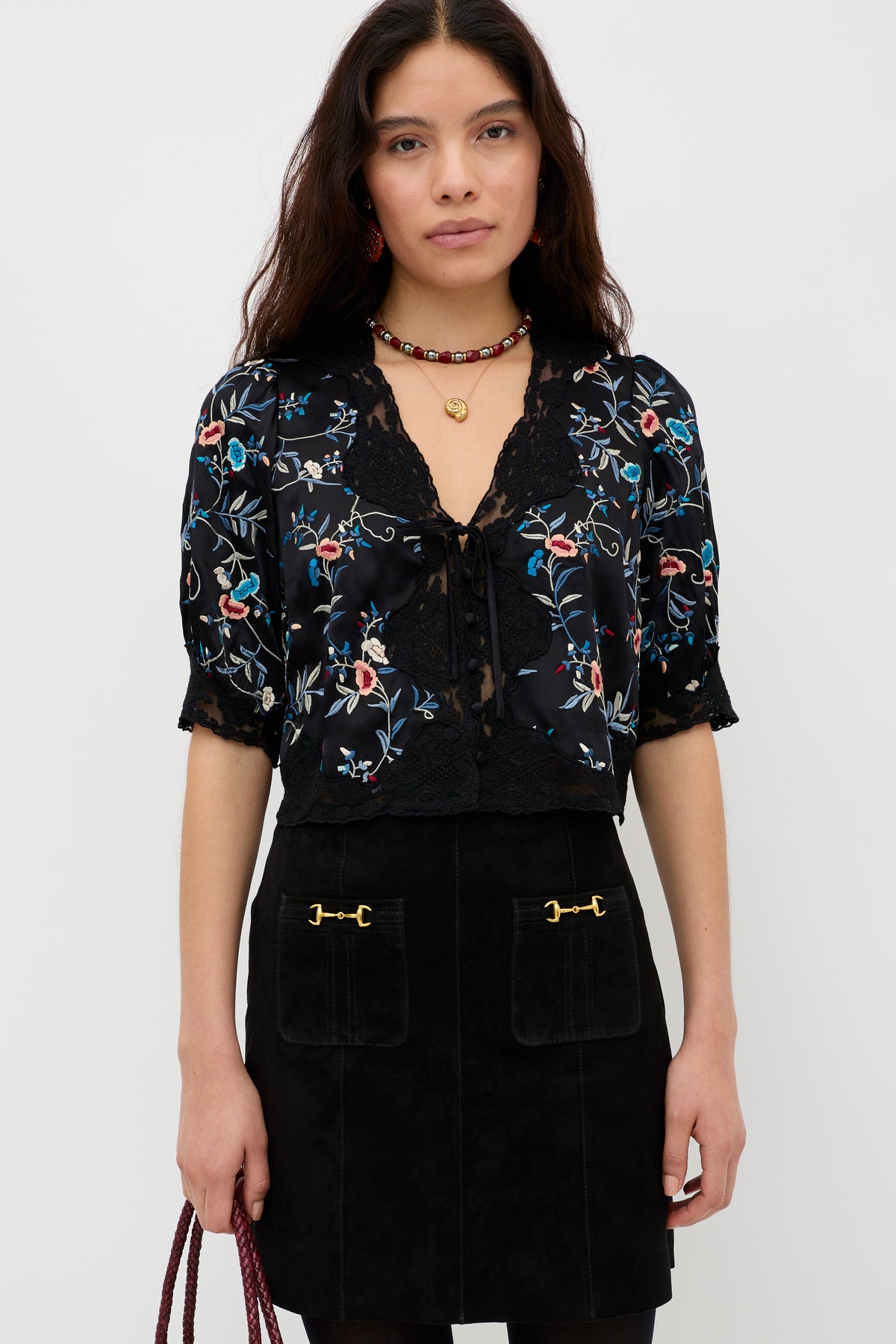Wearing the “Ursula - Botanical Embroidery Black” blouse, a woman with long dark hair pairs it with a black skirt featuring gold details, accessorizes with a beaded necklace, and carries a red bag as she stands against a plain white background.