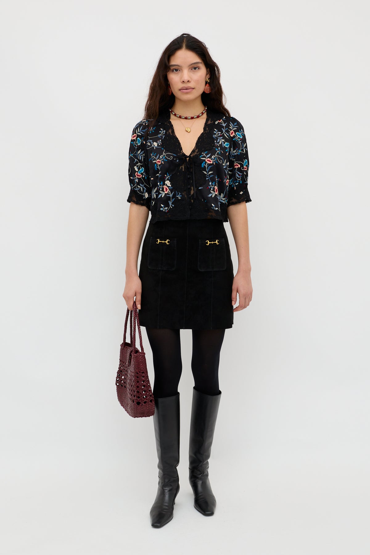 A woman with long dark hair stands against a white background wearing the Ursula - Botanical Embroidery Black blouse, paired with a black mini skirt, black tights, knee-high black boots, a burgundy handbag, and jewelry.