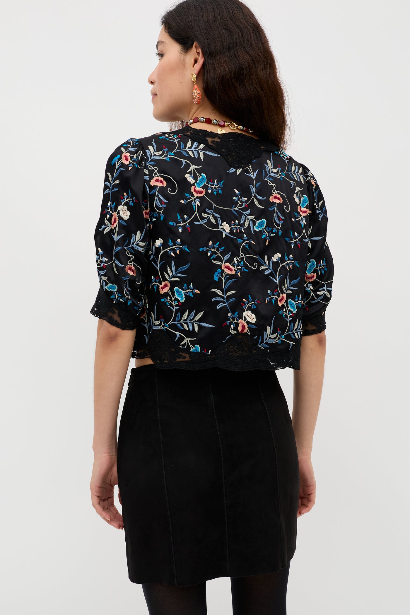 A woman stands facing away, wearing the Ursula - Botanical Embroidery Black crop top paired with a black skirt. She has long brown hair, earrings, and a necklace. Satin lace trim adds an elegant touch to her ensemble, while the plain light-colored background keeps the focus on her vintage-inspired look.