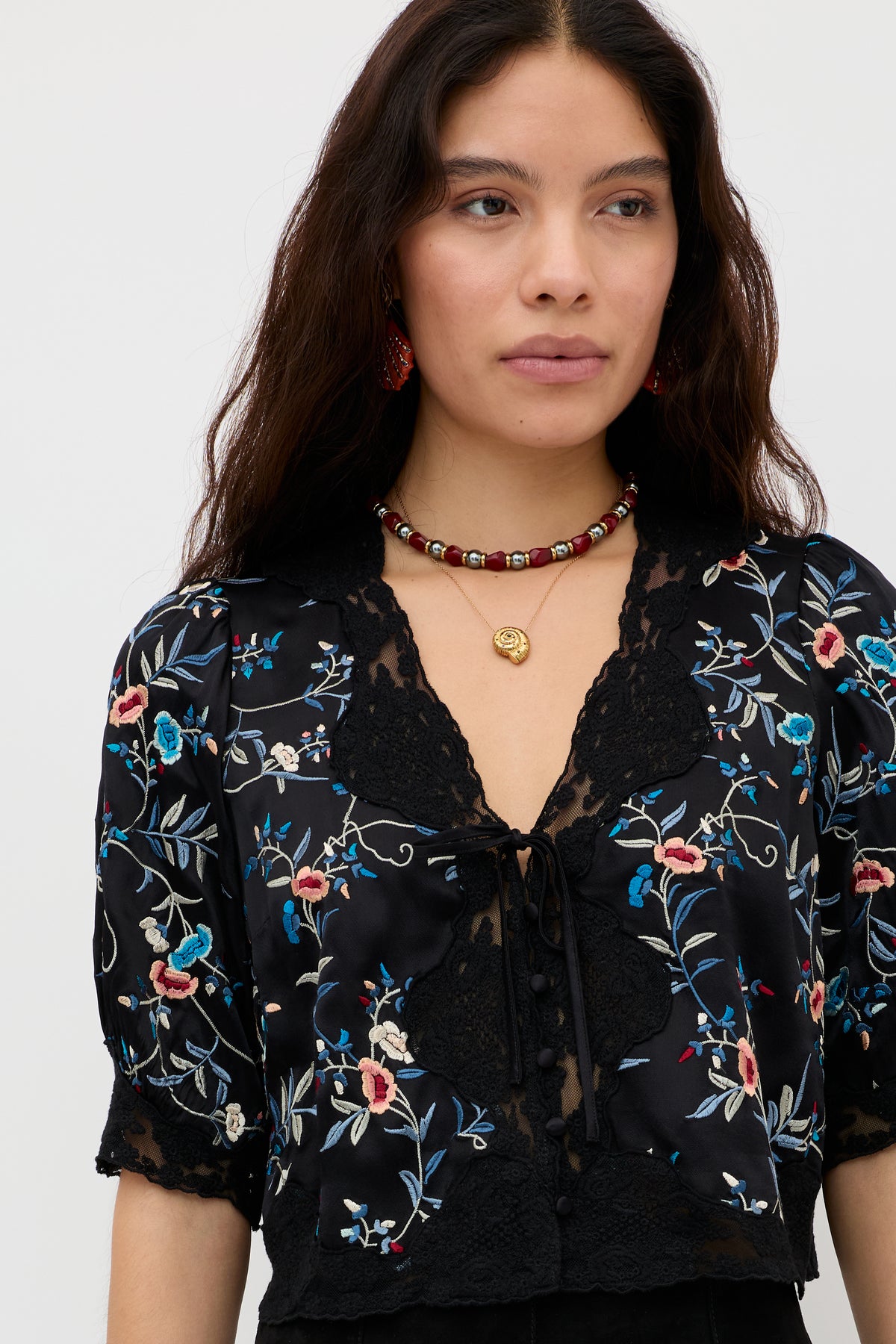 Model is wearing the “Ursula - Botanical Embroidery Black” blouse, featuring silky satin fabric with botanical embroidery and delicate lace accents. She has long dark hair, and her look is complemented by a beaded necklace and red earrings. She stands against a plain white background.