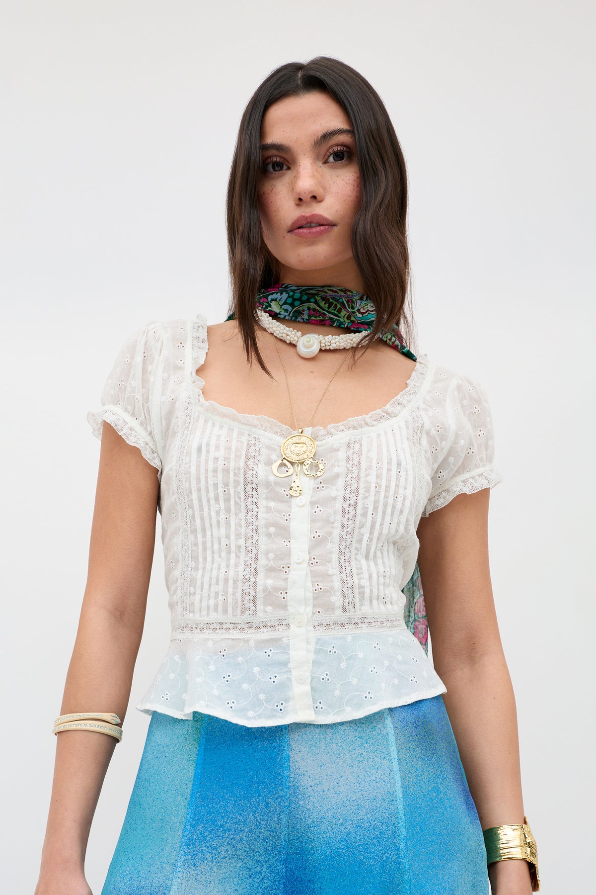 A woman wears the Shereen - Cream sheer eyelet blouse with short puff sleeves, blue ombré pants, layered gold jewelry, and a colorful neck scarf, standing against a light background.