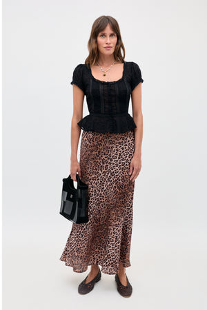 A woman is shown wearing a black short-sleeve blouse paired with the Kelly - Leopard Brown skirt, crafted from luxurious silk crepe de chine. She holds a black handbag, wears dark shoes and a necklace, and stands against a plain white background.