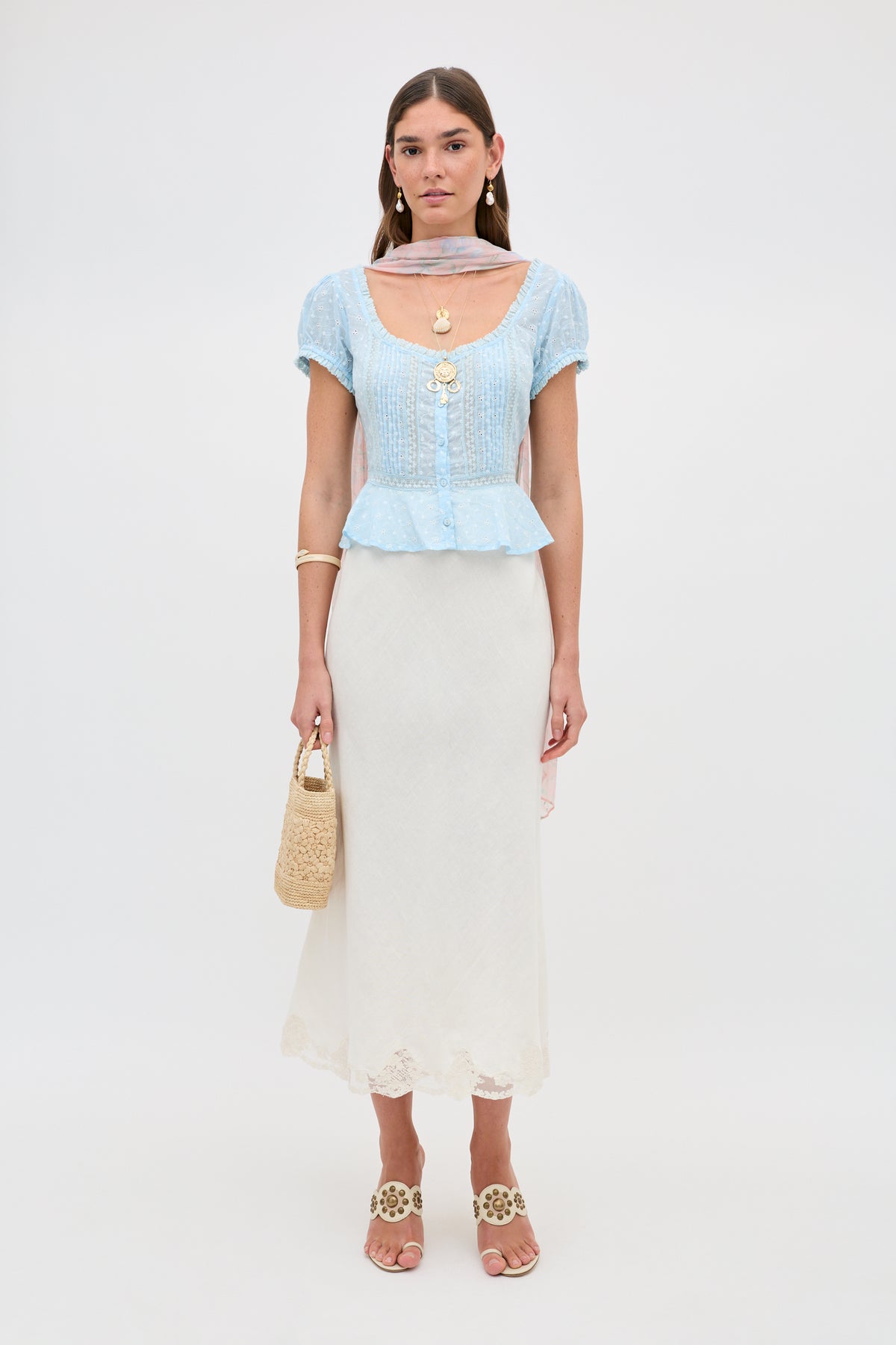 A woman stands against a plain background wearing Sandi - Powder Blue, a peplum hem top paired with a white lace-insert midi skirt, tan sandals, layered necklaces, and holding a woven handbag.