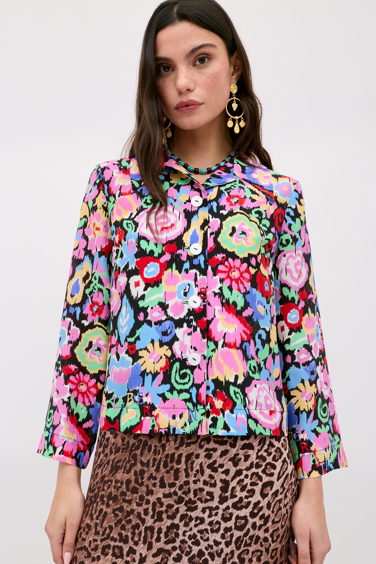 A woman wears the Ginette - Ikat Bloom Charcoal, a colorful floral-patterned button-up blouse with long sleeves, paired with a brown leopard-print skirt. She has long dark hair, gold statement earrings, and stands against a plain light background.