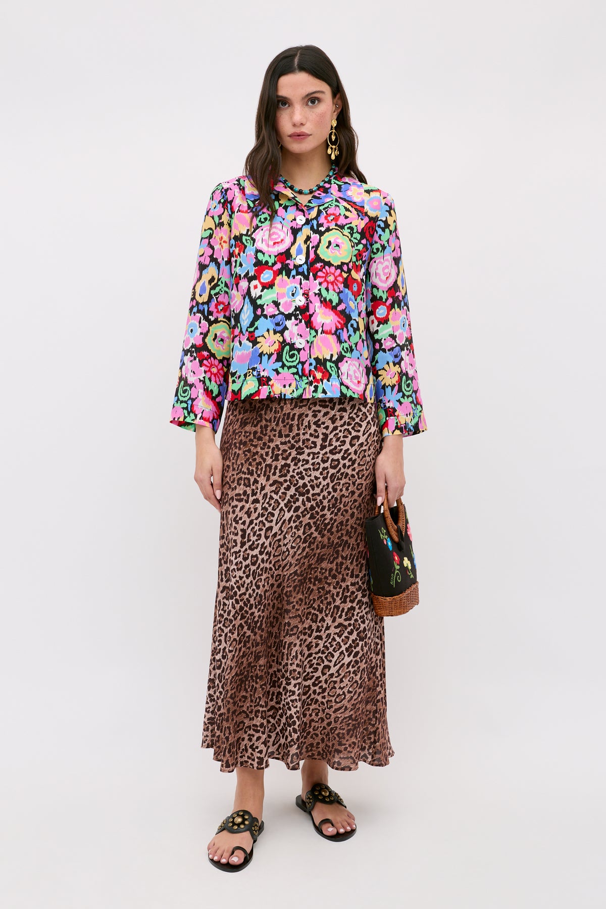 A woman with long dark hair and a neutral expression stands against a plain background wearing the Ginette - Ikat Bloom Charcoal floral long-sleeve top, a leopard print midi skirt, black sandals, and holding a small handbag.