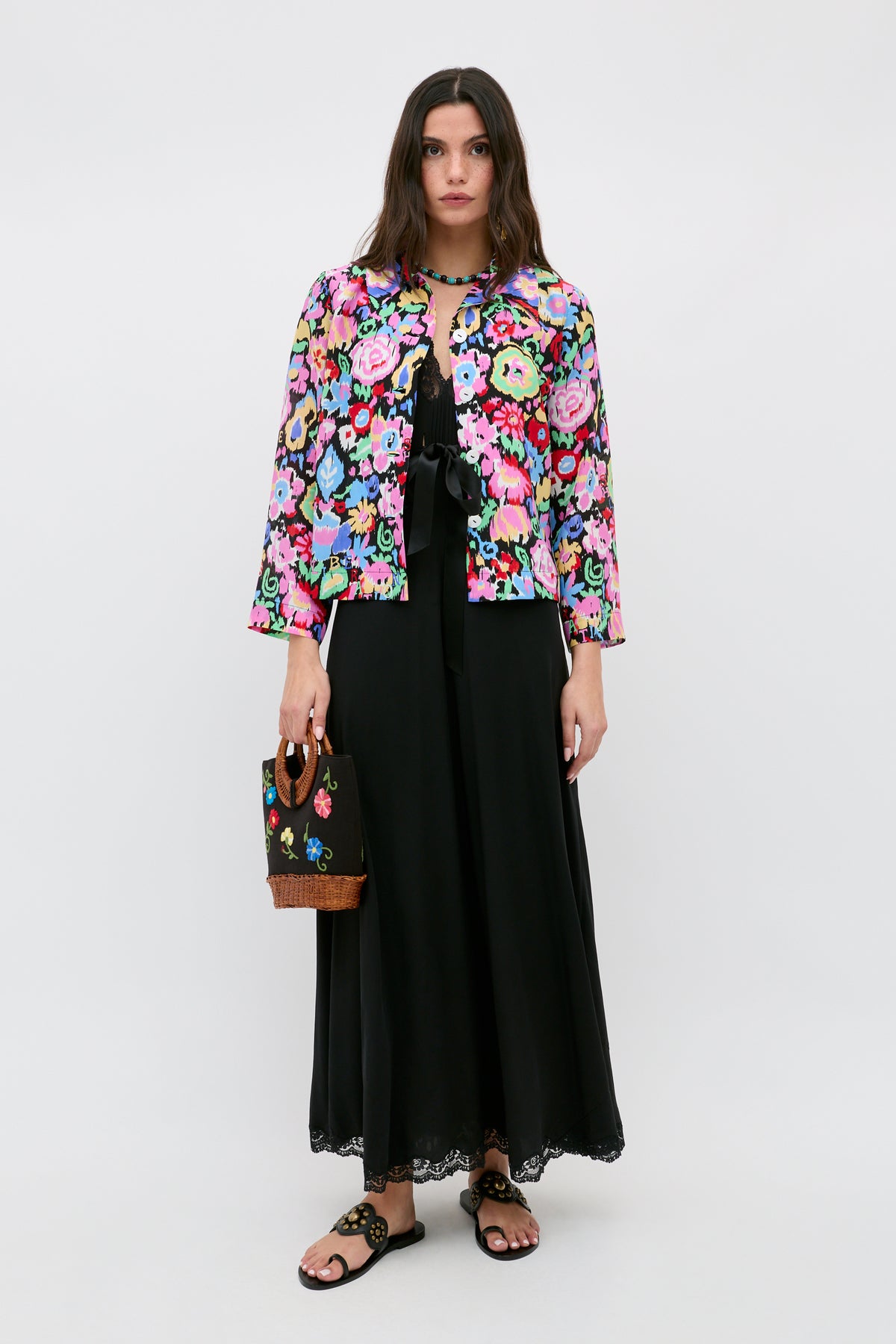 A woman stands against a plain background wearing the Ginette - Ikat Bloom Charcoal, a long black dress, paired with a colorful floral jacket, black sandals, and holding a small black handbag with floral embroidery.