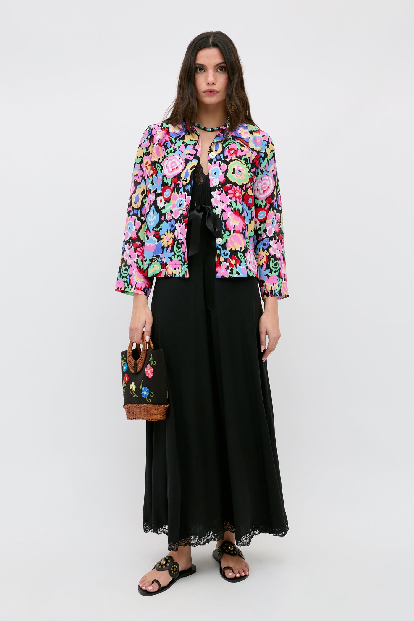 A woman stands against a plain background wearing the Ginette - Ikat Bloom Charcoal, a long black dress, paired with a colorful floral jacket, black sandals, and holding a small black handbag with floral embroidery.