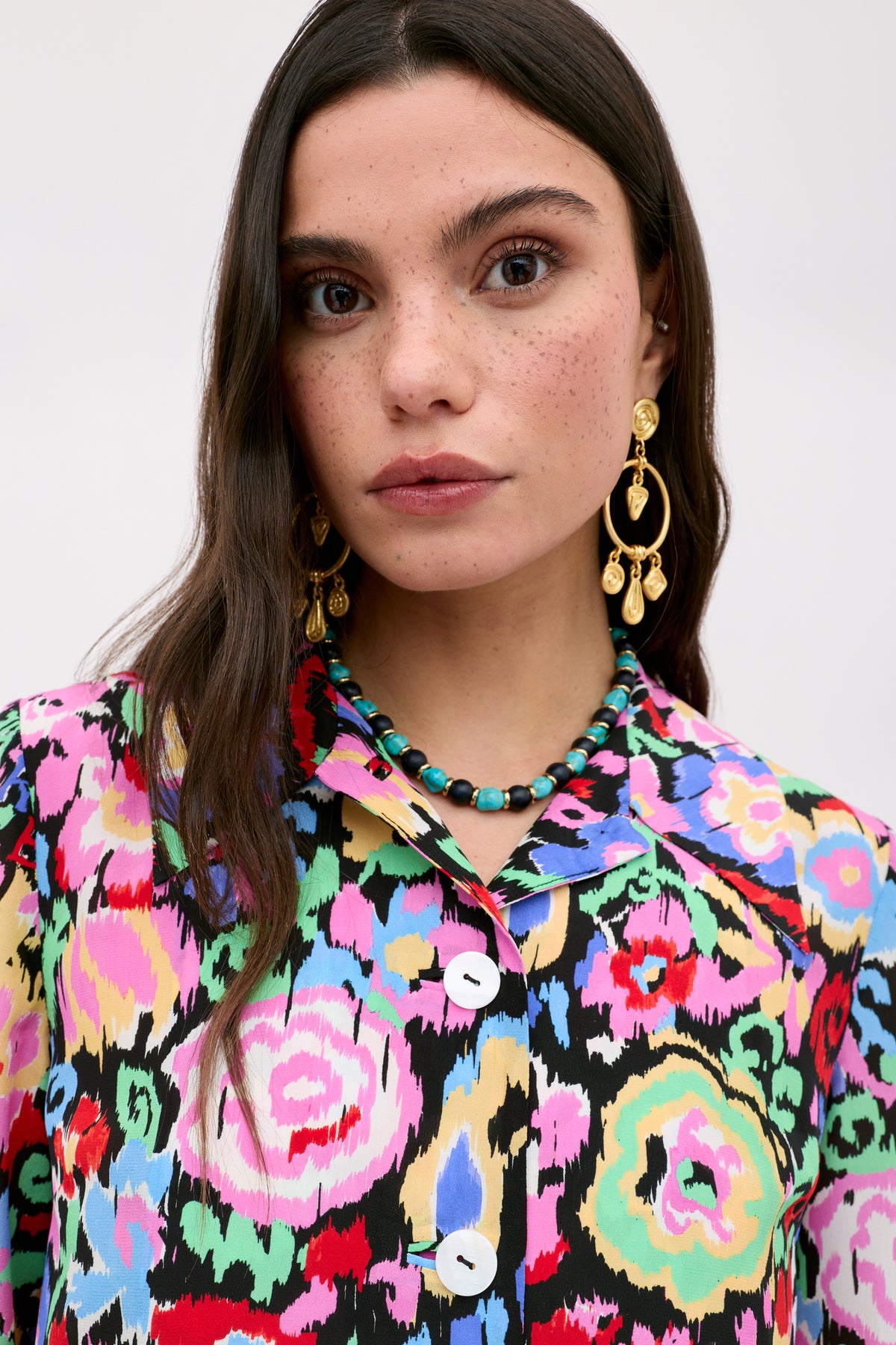 A woman with long brown hair and freckles models the Ginette - Ikat Bloom Charcoal floral shirt with large buttons. She wears gold statement earrings and a turquoise beaded necklace, looking directly at the camera against a plain background.