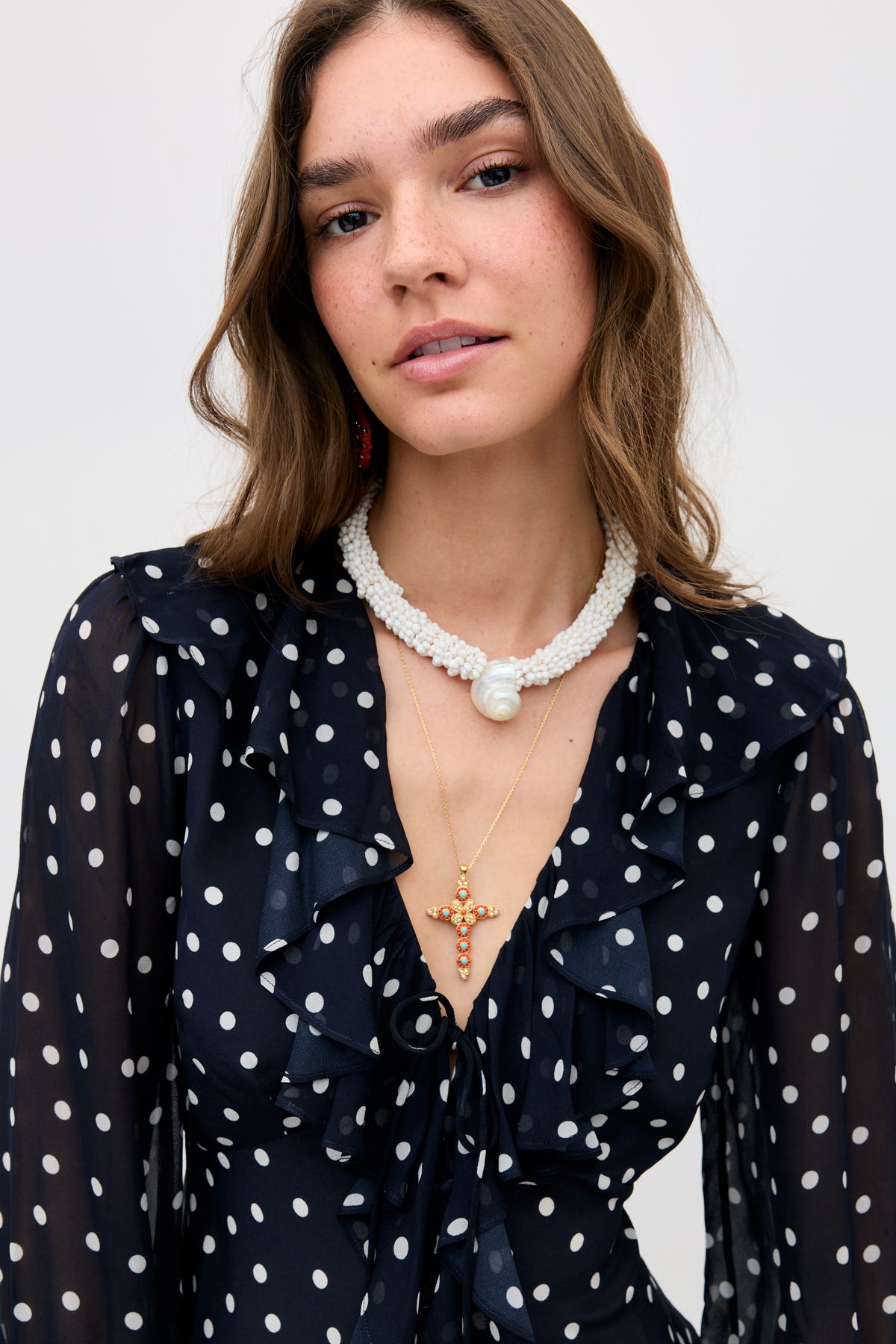 Woman with long brown hair wearing the Lenka - Gold necklace, a navy polka dot ruffled blouse, and a white beaded necklace with a large pendant, posing against a plain background.