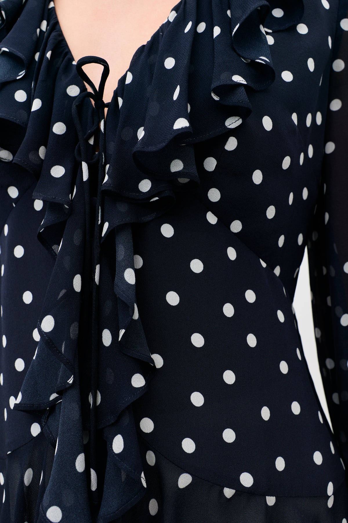 Close-up of the Ami - Layla Dot Navy blouse in dark blue with white polka dots, sheer lightweight fabric, ruffled neckline and front, and a thin black tie at the neck.
