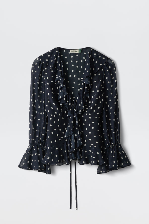 The Ami - Layla Dot Navy is a navy blue long-sleeve blouse featuring white polka dots, a ruffled neckline and cuffs, and a waist tie, shown against a plain white background.
