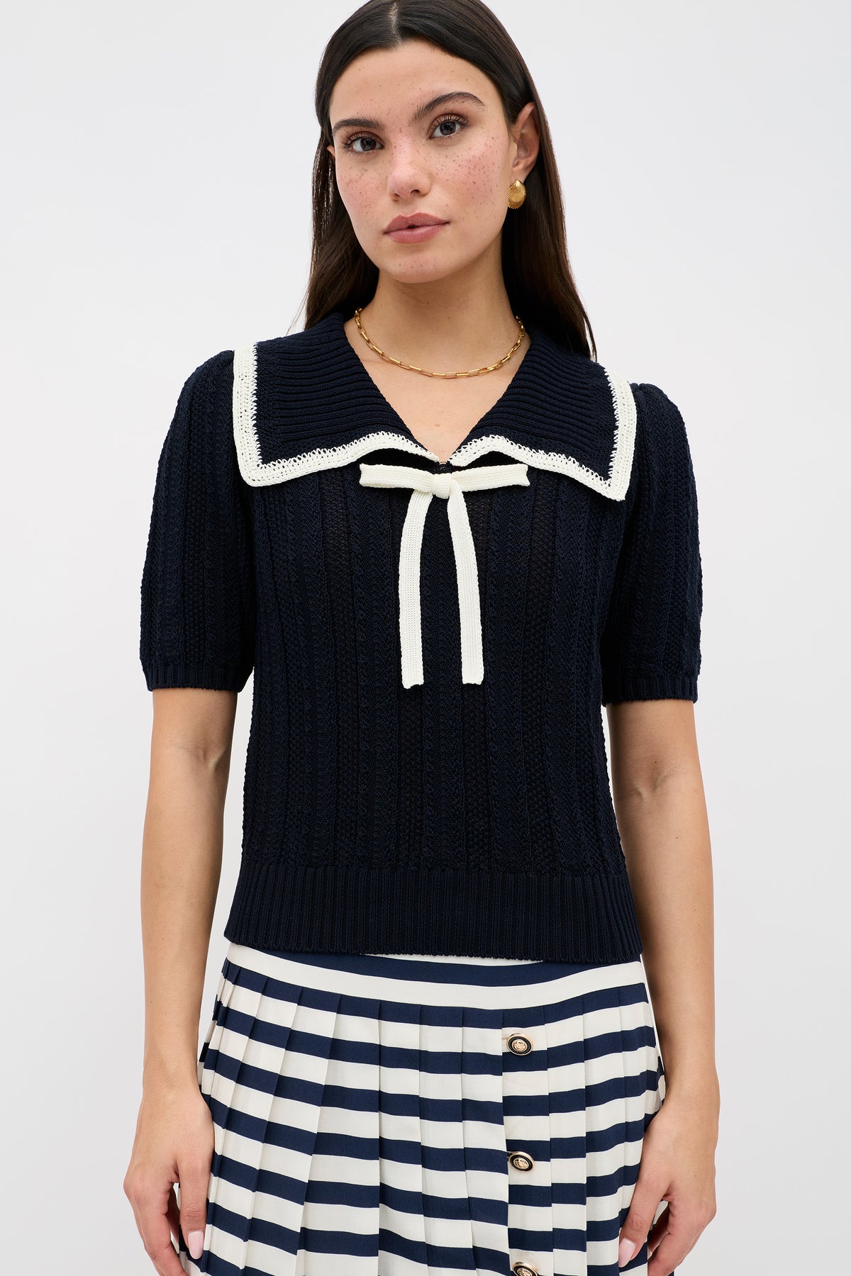 A woman stands against a plain background wearing the 'Pippa - Deep Navy' knit top with a sailor collar and white bow, paired with a navy and white striped pleated skirt featuring buttons.