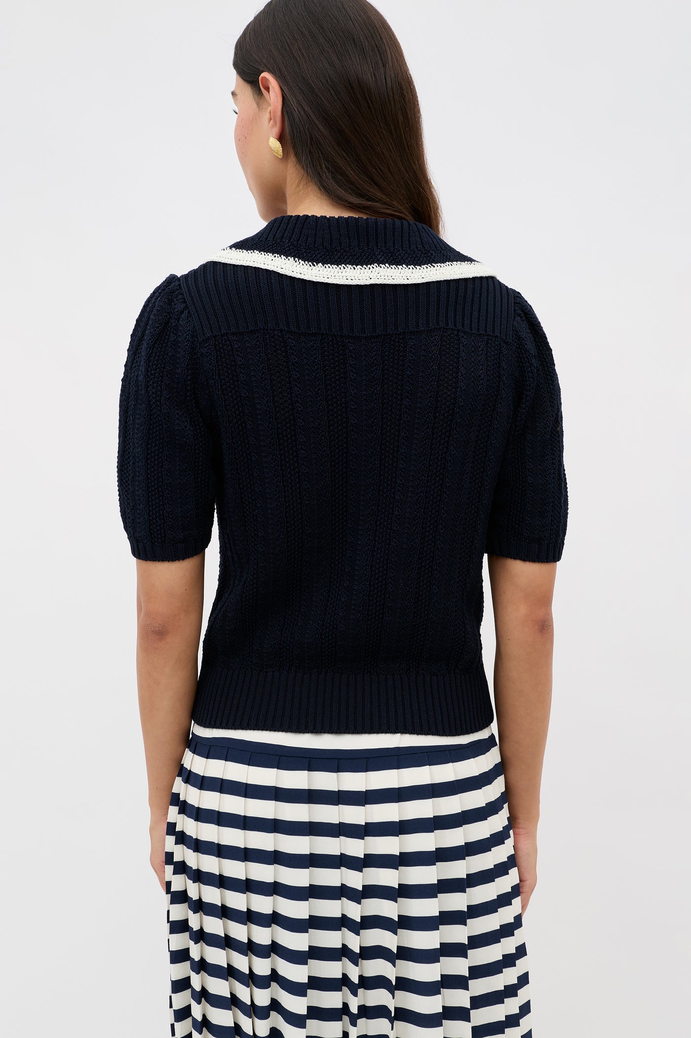 A woman with long brown hair, seen from behind, wears the Pippa - Deep Navy short-sleeve knit top with a white-collared accent and a pleated skirt featuring navy and white horizontal stripes.