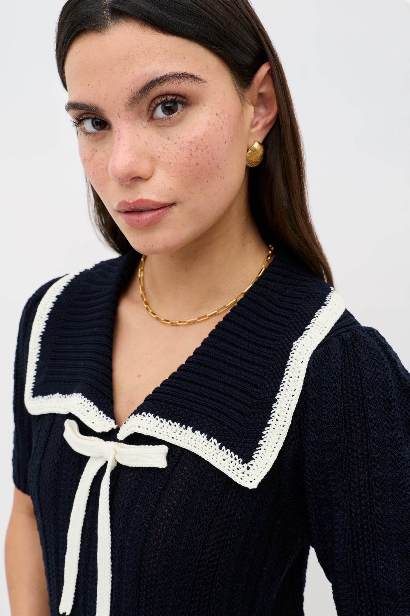 A woman with long dark hair wears gold earrings, a gold chain necklace, and the Pippa - Deep Navy knit top featuring a large sailor collar with white trim and a white bow. She has freckles and looks directly at the camera.