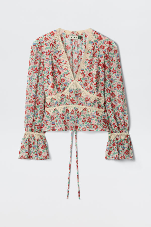The Lenara - Jeannie Bloom Ivory is a long-sleeved, v-neck blouse featuring a red and blue floral print, lace trim, flared cuffs, and a waist tie, shown against a plain white background.
