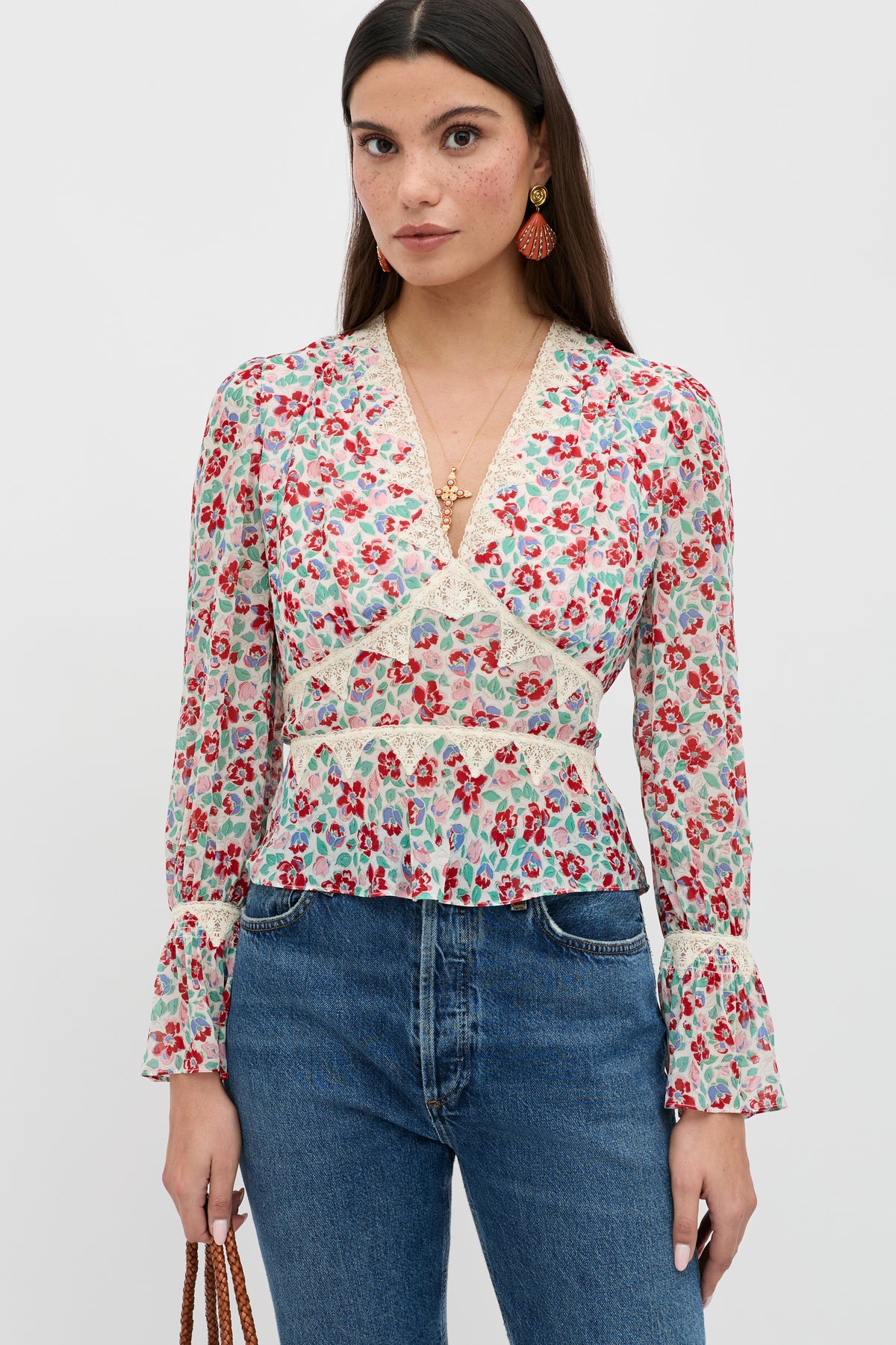 A woman with long dark hair models the Lenara - Jeannie Bloom Ivory floral V-neck blouse with ruffled cuffs, styled with blue jeans, a brown bag, statement earrings, and a gold necklace against a plain white background.