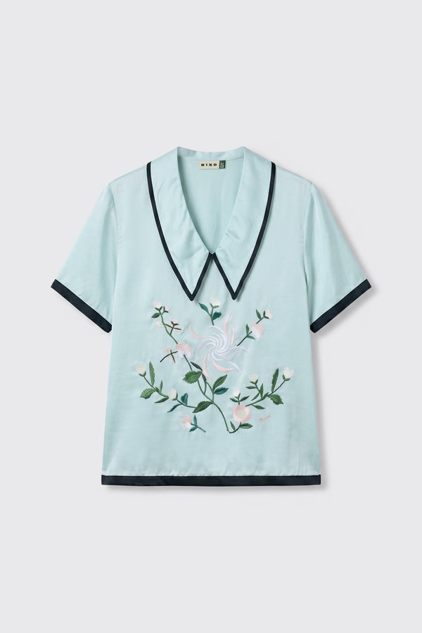 Nicolina - Frida Embroidery Mist is a light blue short-sleeve blouse featuring a wide pointed collar, black trim, and pink floral embroidery on the front. The blouse is displayed laid flat on a plain white background.