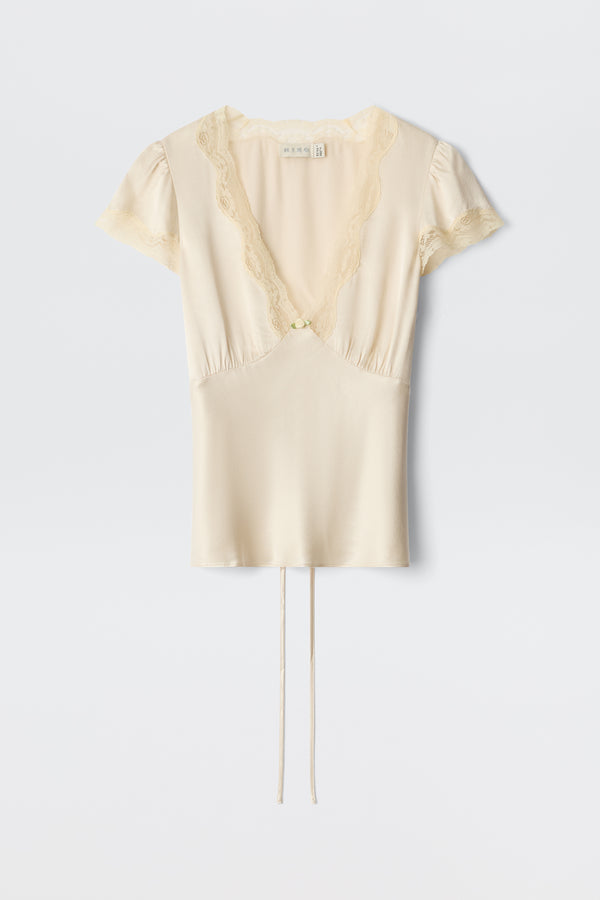 The Karia – Champagne is a cream-colored silk blouse featuring short sleeves, lace-trimmed deep V-neckline and sleeves, a center button, and two thin ties at the hem, shown against a plain white background.
