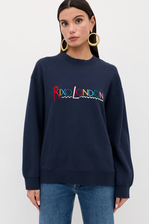 A woman in the Blaze - London Embroidery Navy sweatshirt stands facing forward, wearing gold hoop earrings and blue jeans. She has straight dark hair and poses against a plain white background.