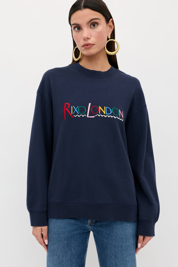 A woman in the Blaze - London Embroidery Navy sweatshirt stands facing forward, wearing gold hoop earrings and blue jeans. She has straight dark hair and poses against a plain white background.