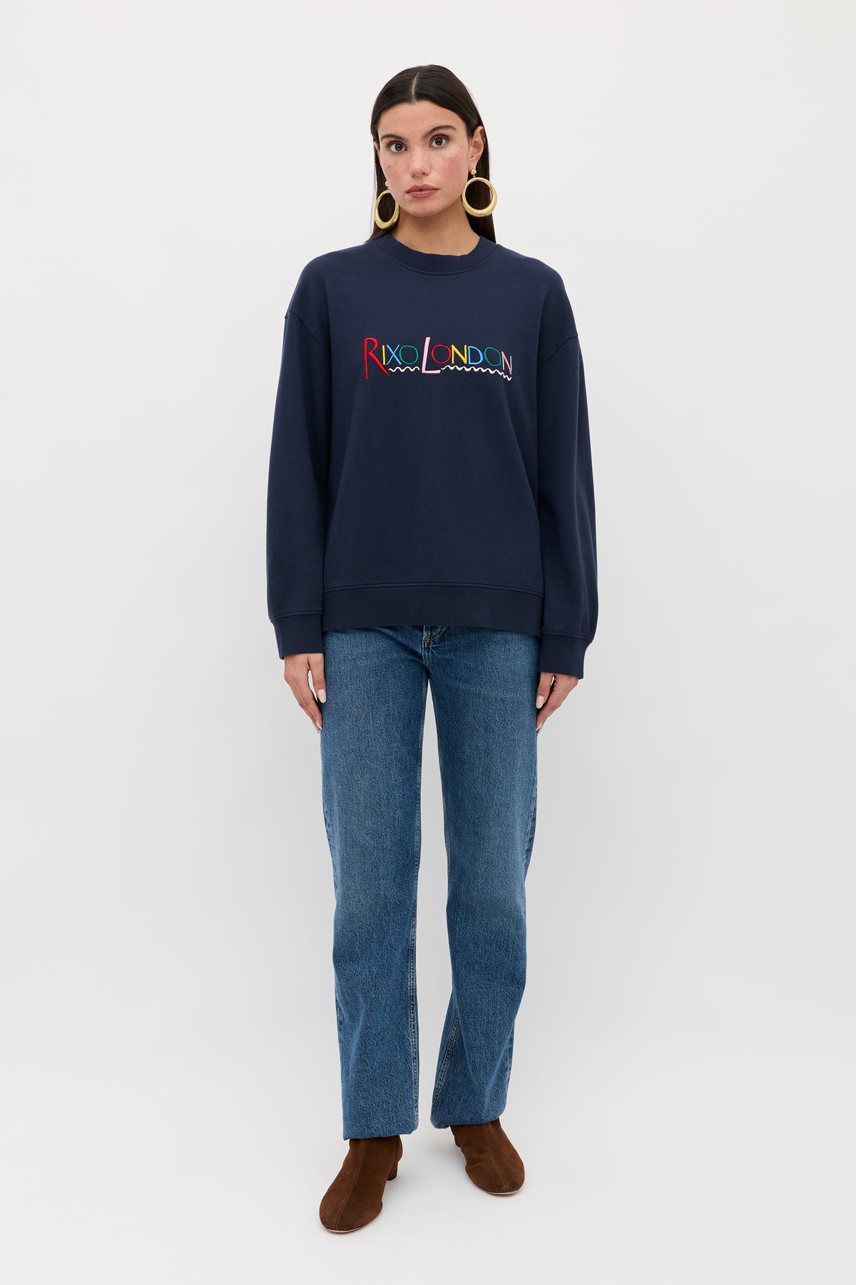 A woman stands against a plain white background wearing the Blaze - London Embroidery Navy sweatshirt from RIXO, paired with blue jeans, brown boots, and large hoop earrings. She faces forward with arms relaxed at her sides.