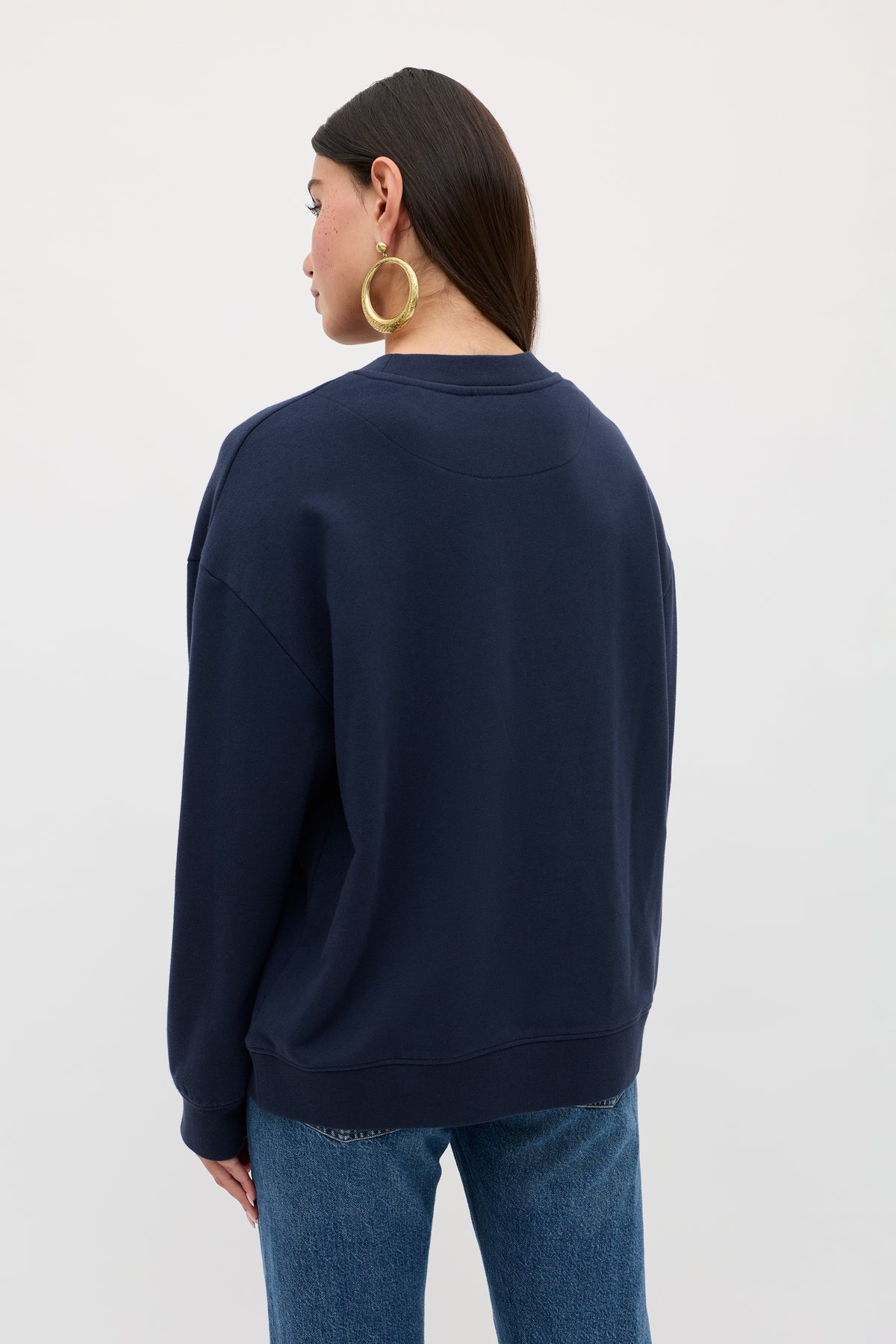 A person with long dark hair, gold hoop earrings, and blue jeans stands with their back to the camera against a plain light background, wearing the Blaze - London Embroidery Navy sweatshirt.