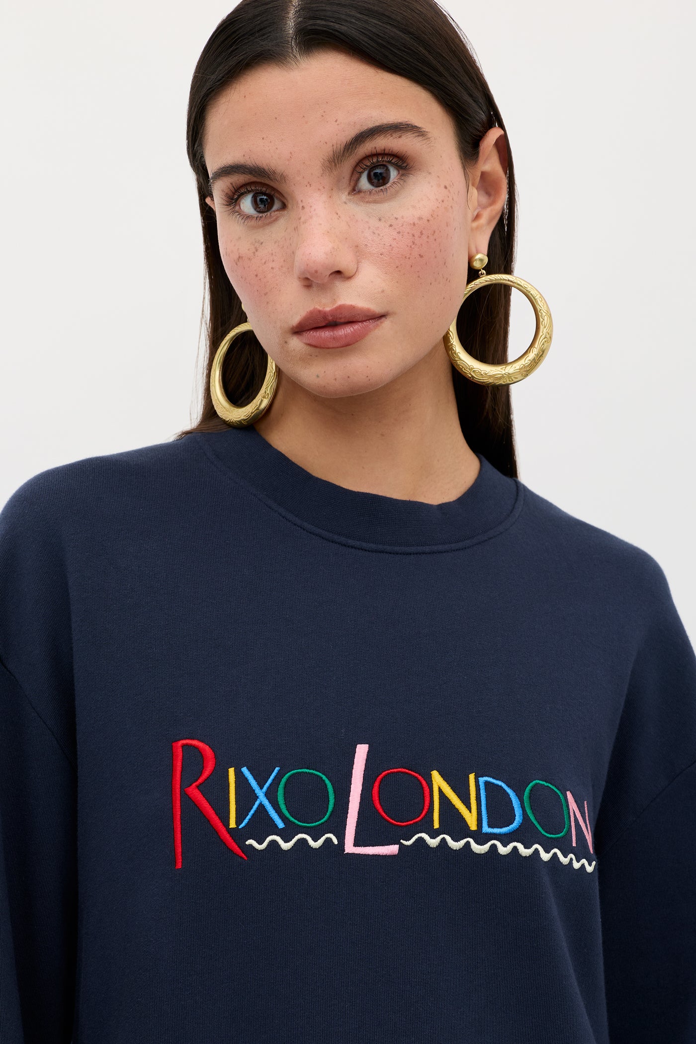 A woman with long dark hair, freckled skin, and gold hoop earrings wears the Blaze - London Embroidery Navy sweatshirt. "London" is embroidered in colorful letters. She stands against a simple light background.