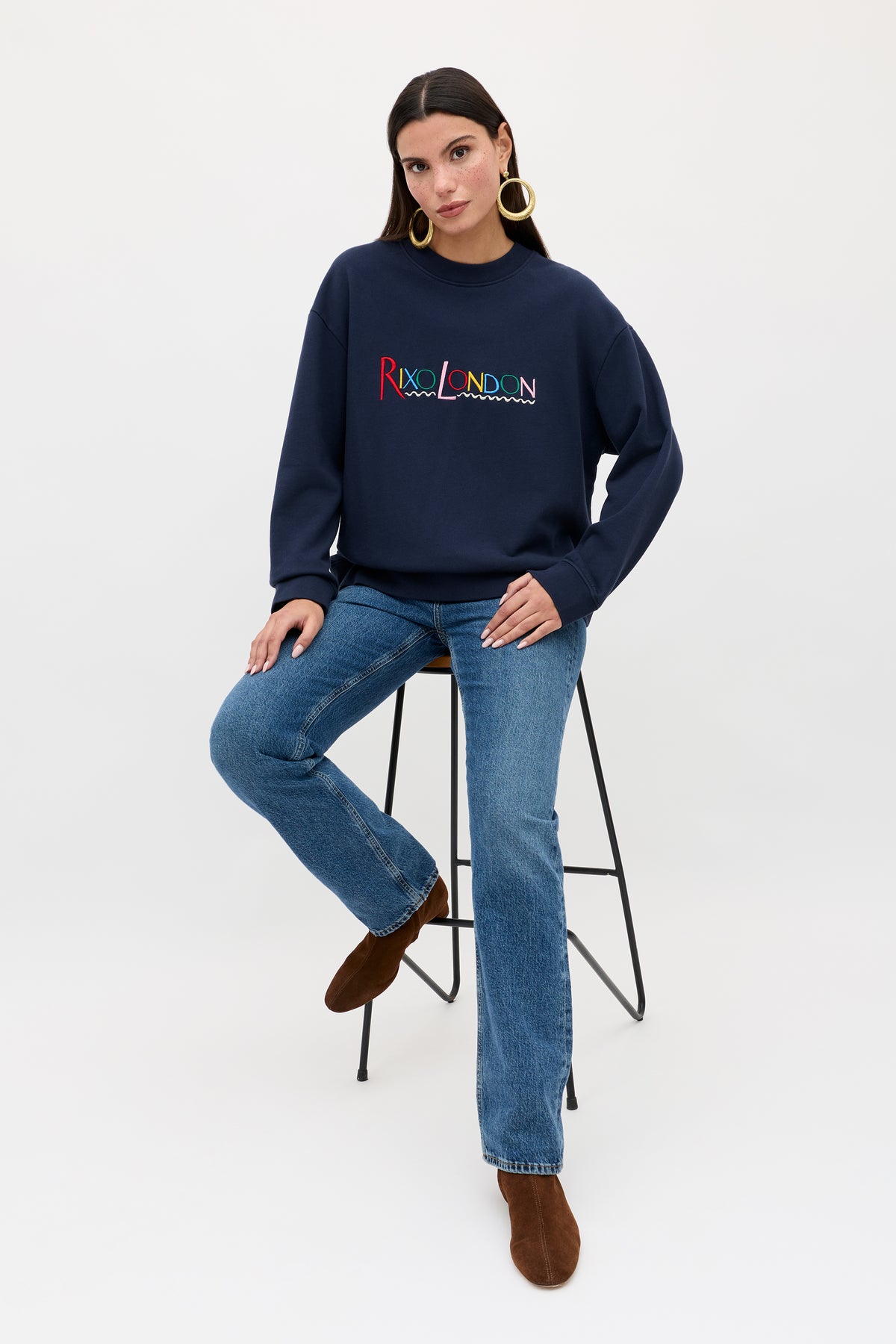 A woman with straight dark hair sits on a tall stool against a white background wearing the Blaze - London Embroidery Navy sweatshirt, blue jeans, brown boots, and large gold hoop earrings.