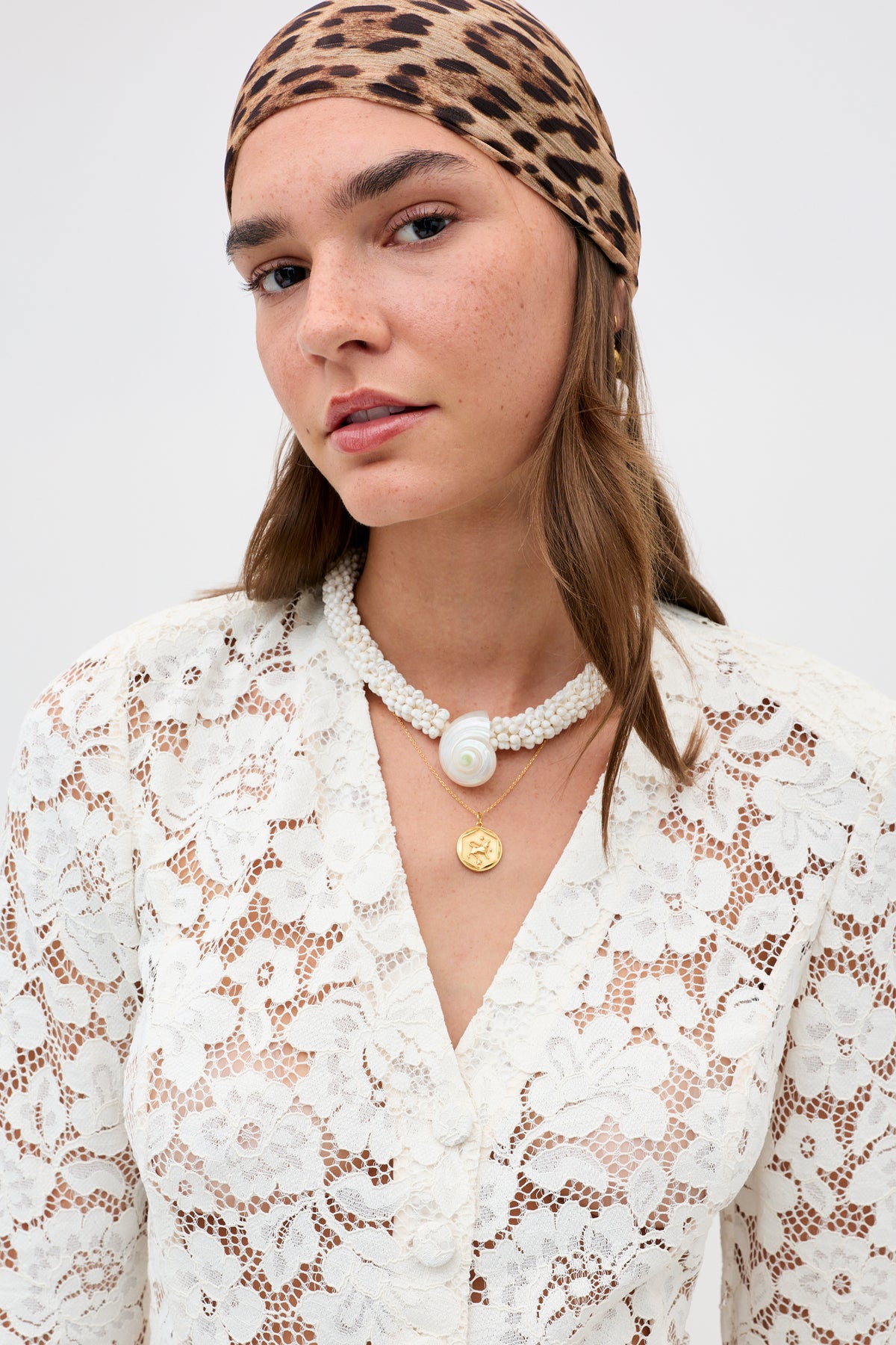 A woman models the Imara - Pearl necklace, featuring pearls and a large shell pendant, paired with a leopard-print headscarf, a white lace top, and a gold necklace against a plain background.