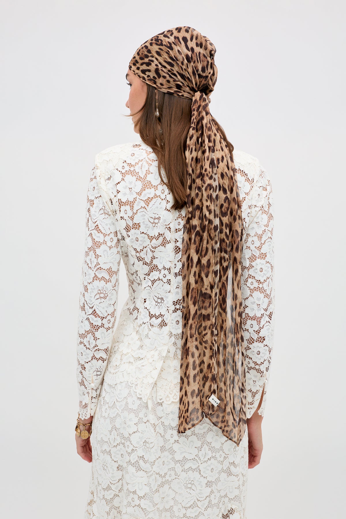 A woman in a long-sleeved white lace dress and straight brown hair faces away, wearing the Padma - Cream headscarf with a leopard print, draped down her back against a plain white background.