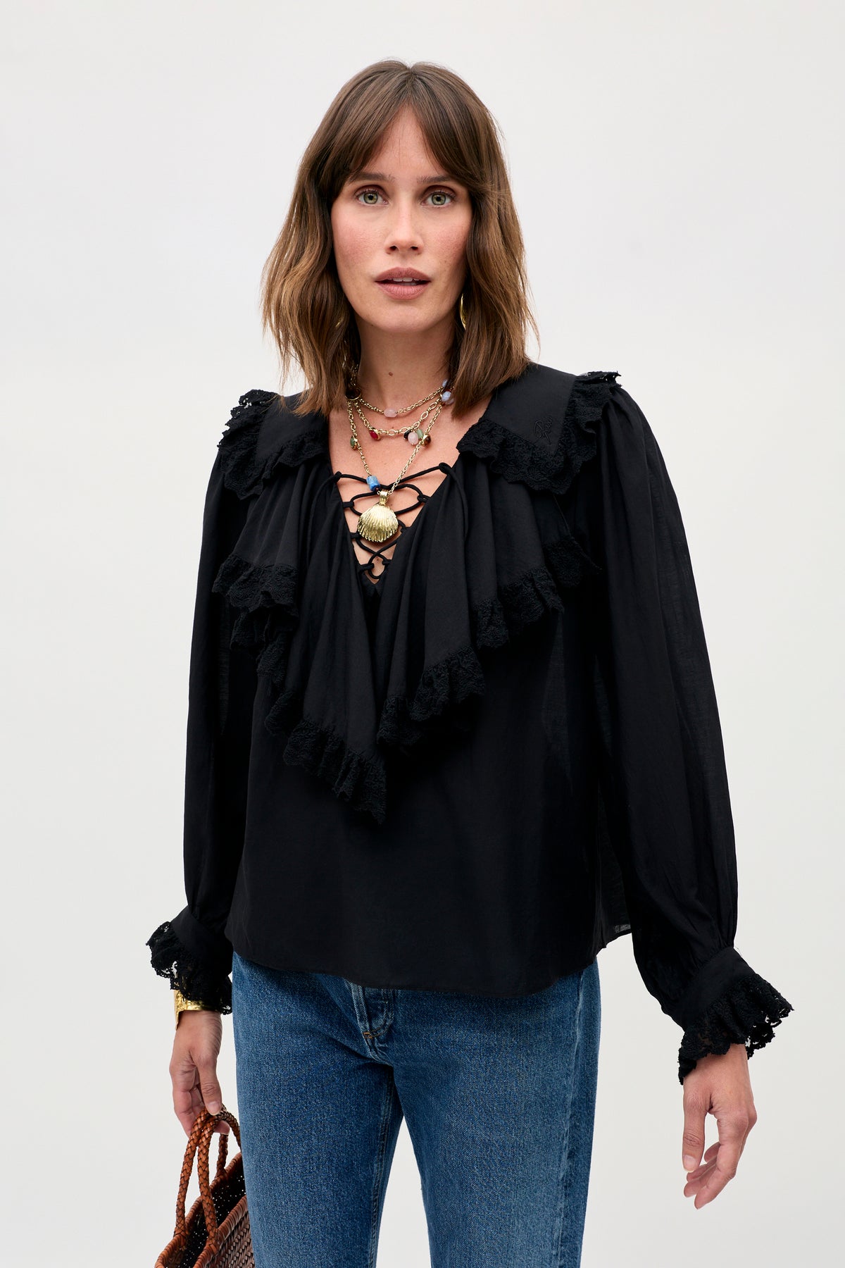 A woman with shoulder-length brown hair stands against a plain light background wearing the Milia - Black ruffled blouse with lace-up front detail, paired with blue jeans, layered necklaces, and holding a brown woven bag.