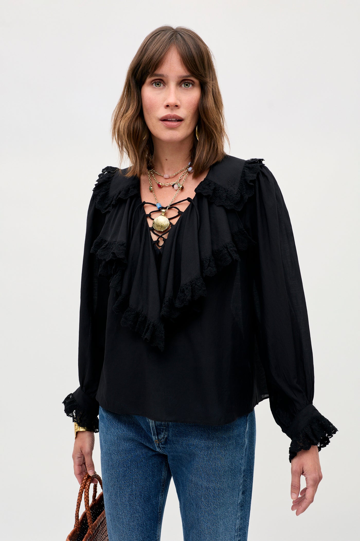 A woman with shoulder-length brown hair stands against a plain light background wearing the Milia - Black ruffled blouse with lace-up front detail, paired with blue jeans, layered necklaces, and holding a brown woven bag.