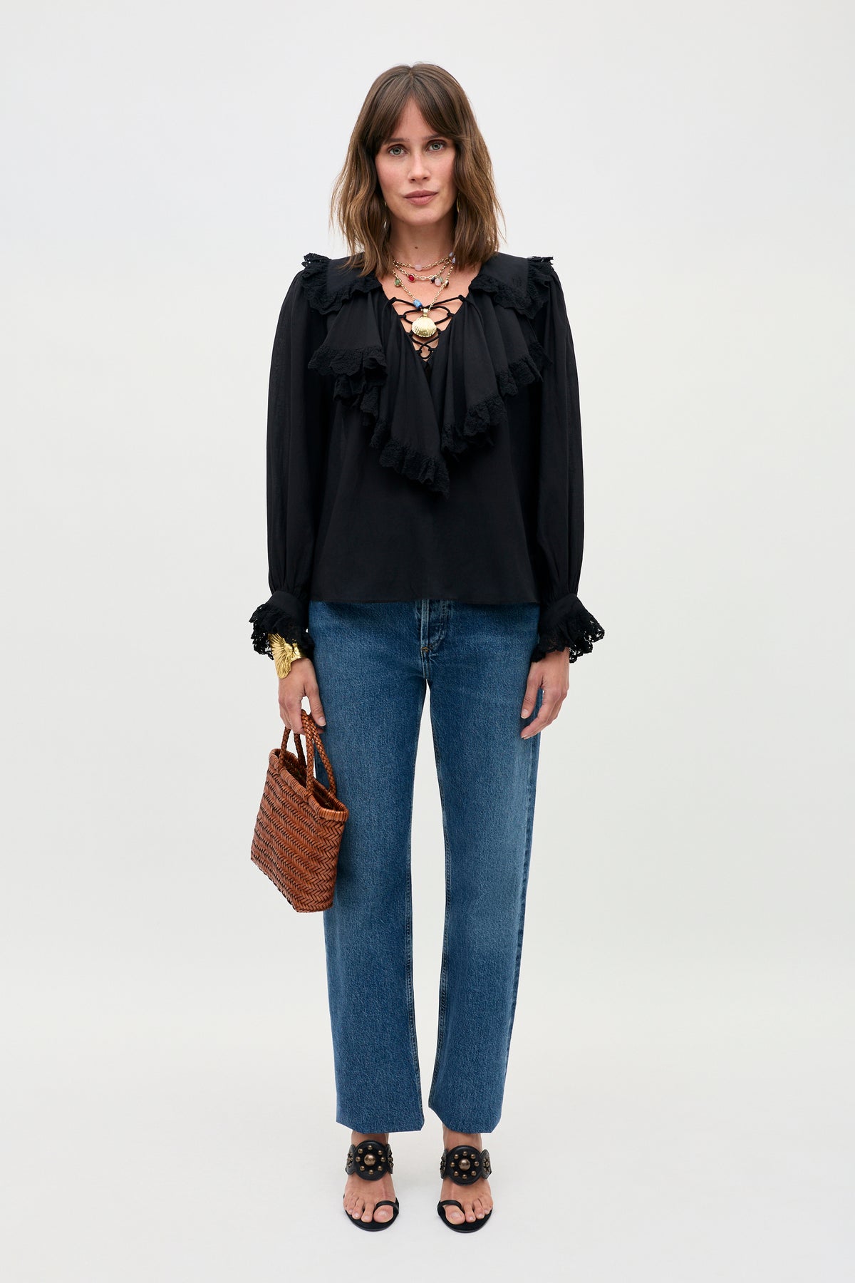 A woman stands against a plain background wearing the Milia - Black ruffled blouse, blue jeans, black sandals, a chunky necklace, and holding a small woven brown handbag.