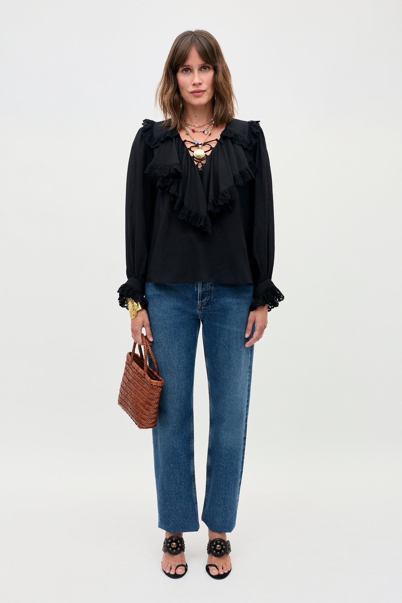 A woman stands against a plain background wearing the Milia - Black ruffled blouse, blue jeans, black sandals, a chunky necklace, and holding a small woven brown handbag.