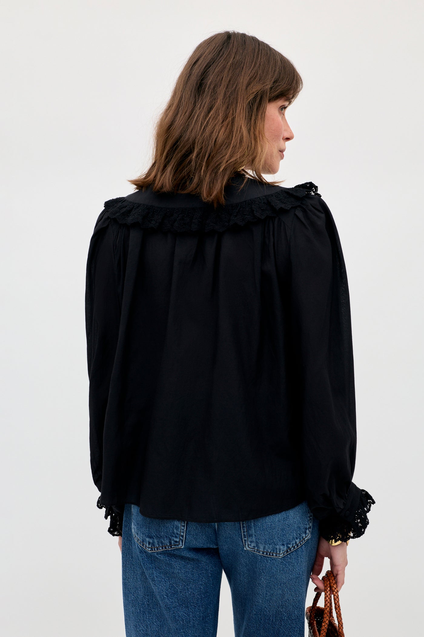 A person with shoulder-length brown hair is wearing the Milia - Black long-sleeve blouse with lace details and blue jeans, standing facing away from the camera against a plain background.