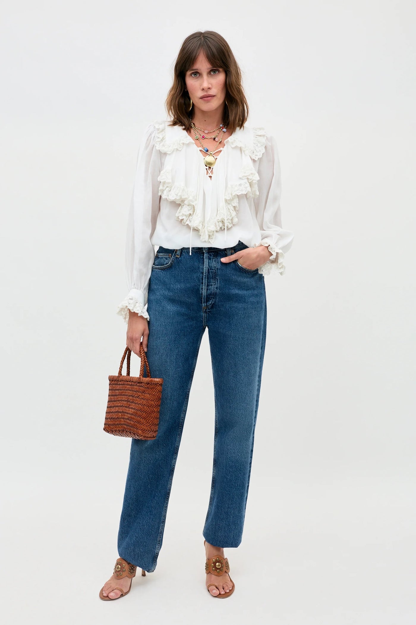 A woman with shoulder-length brown hair wears the Milia - White ruffled blouse, blue jeans, tan heeled sandals, layered necklaces, and holds a small woven handbag against a plain white background.