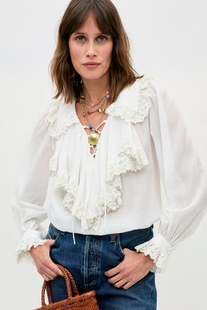 A woman with shoulder-length brown hair stands against a plain background, wearing the Milia - White blouse with lace ruffles and blue jeans. She holds a woven bag and accessorizes with layered necklaces.