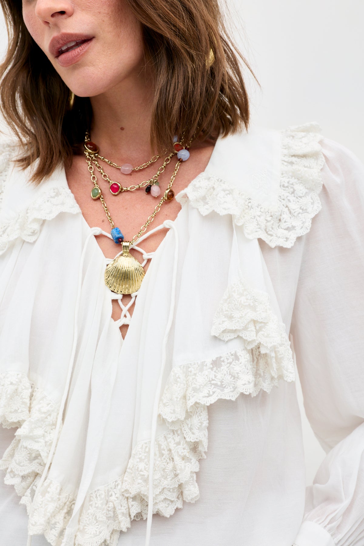A woman wearing a white ruffled blouse with lace trim and layered necklaces, featuring the Lima - Gold shell pendant along with colorful beads. Only her lower face, neck, and upper torso are visible.